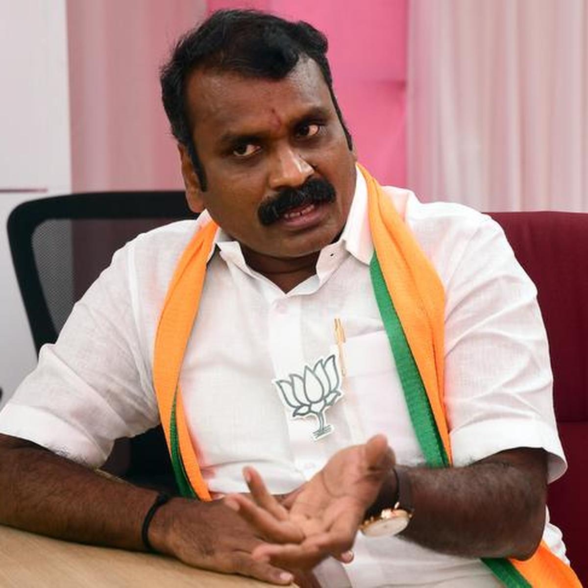 Dr. L. Murugan, in a white shirt with a party scarf, seated at a table and speaking with an open hand gesture