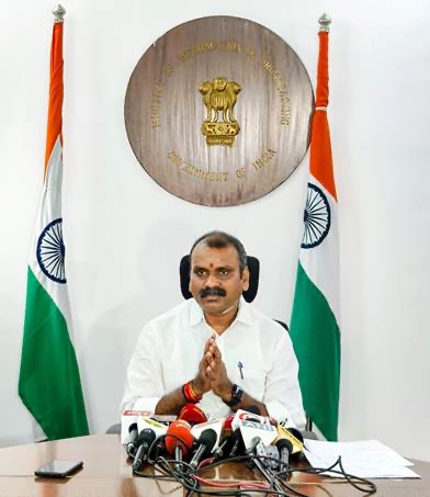 Dr. L. Murugan, hands folded in greeting, seated at a table with a bank of microphones, in front of the Ministry of Information & Broadcasting emblem and two national flags