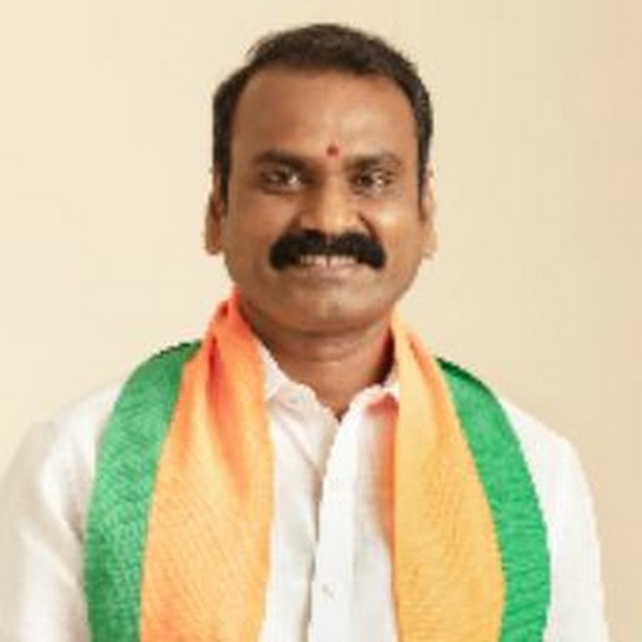 Head-and-shoulders portrait of Dr. L. Murugan, smiling, wearing a white shirt with a saffron and green scarf, against a cream background