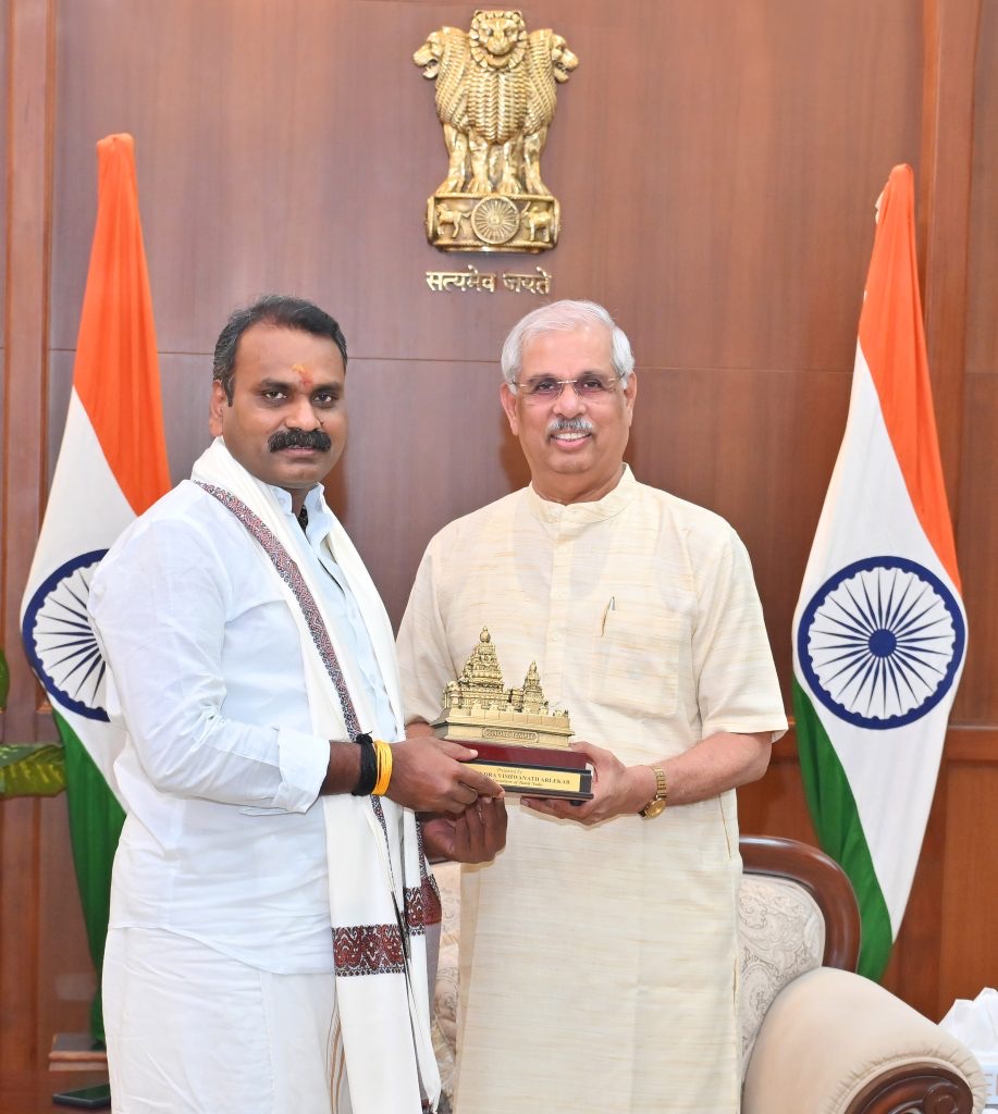 Dr. L. Murugan, wearing a white shirt and a woven stole, receives a gilded temple-shaped memento from a dignitary in a hall with the State Emblem and two national flags