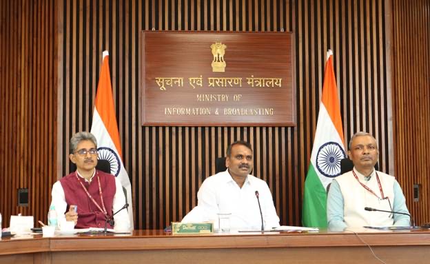 Dr. L. Murugan seated at the centre of a conference table between two officials, beneath the Ministry of Information & Broadcasting nameboard and two national flags