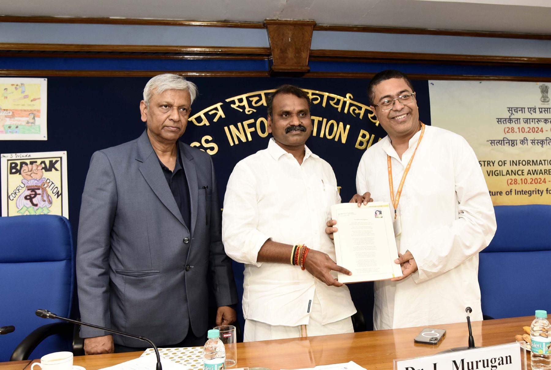 Dr. L. Murugan, standing between two officials, holds a document at a programme in the Press Information Bureau hall, with the PIB name in gold letters behind them