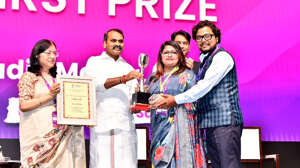 Dr. L. Murugan hands a microphone-shaped trophy to winners on stage at the National Community Radio Awards; a framed First Prize certificate is held beside him