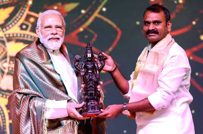 On a stage with a painted backdrop, Dr. L. Murugan presents a dark carved deity sculpture to Prime Minister Narendra Modi, who wears a patterned shawl