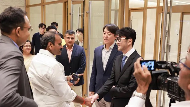 Dr. L. Murugan shakes hands with a group of executives in suits in a glass-walled lobby while a camera operator records the meeting