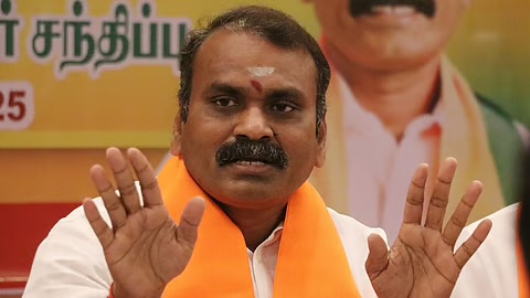 Dr. L. Murugan, wearing a saffron scarf, gestures with both hands raised while speaking at a press meet