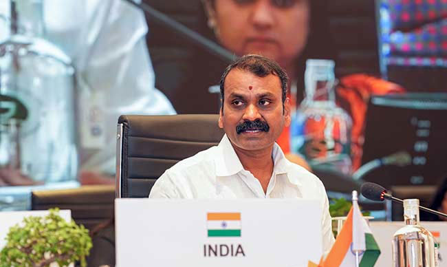 Dr. L. Murugan seated at a conference table behind a nameplate reading INDIA, with a small national flag and a microphone