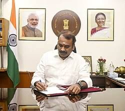 Dr. L. Murugan signing a file at his desk, with portraits of the Prime Minister and the President on the wall behind