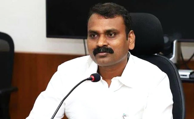 Dr. L. Murugan, in a white shirt, seated at a meeting with a microphone in front of him