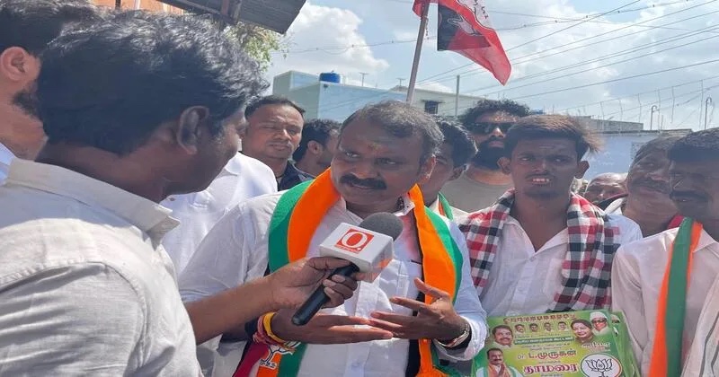 Dr. L. Murugan, wearing a party scarf, speaks into a television microphone while surrounded by supporters in a street
