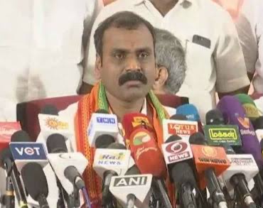 Dr. L. Murugan, wearing a party scarf, addresses a press conference behind a cluster of television channel microphones