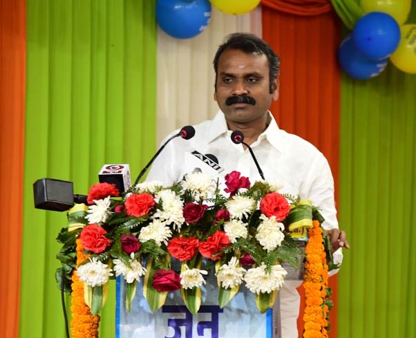 Dr. L. Murugan speaks at a podium decorated with roses and chrysanthemums, with microphones in front of him