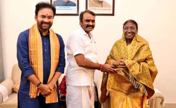 Dr. L. Murugan stands between two guests, clasping the hand of Smt. Droupadi Murmu, who wears a yellow shawl